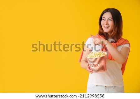 Young Asian woman with tasty popcorn pointing at something on yellow background