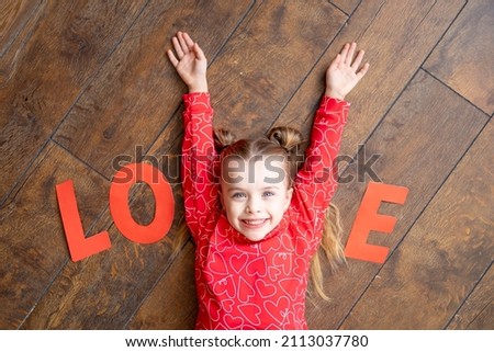 a little girl child in red pajamas with the inscription Love lies on a dark brown wooden background on the floor and laughs holding her hands in the shape of the letter V, Valentine's day