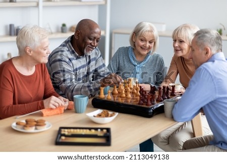 Multiracial senior people men and women in casual playing chess at nursing home, sitting around table, drinking tea with cookies, playing table games, knitting, spending time together Royalty-Free Stock Photo #2113049138