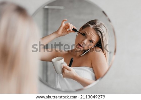 Busy young lady talking on smartphone, applying mascara and drinking coffee simultaneously in a hurry, being late for work, standing near mirror in bathroom Royalty-Free Stock Photo #2113049279