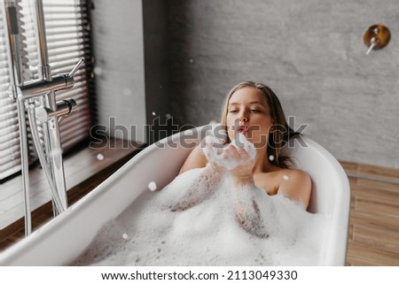 Young woman blowing soap bubbles and having fun while lying in bathtub full of foam at home, copy space. Charming lady relaxing in bath and enjoying beauty and skincare day Royalty-Free Stock Photo #2113049330