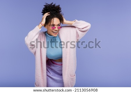 cheerful african american woman in stylish sunglasses adjusting curly hair isolated on violet