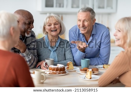 Multiracial group of happy senior people sitting around table drinking tea with cake and having conversation, smiling and laughing, having home party or enjoying time at nursing home Royalty-Free Stock Photo #2113058915