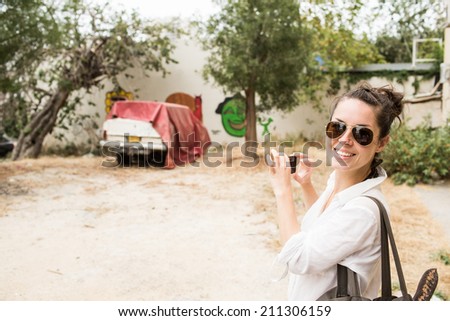 Happy young woman smiling while taking snapshot with cell phone camera the vintage grunge old car in the messy place in the city in Tel Aviv. Unusual moment from vocation or travel.