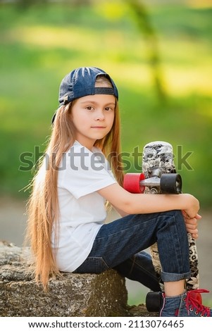 A little girl in a baseball cap, back to front, sits on a stone, holding a skateboard with one hand on her knee. Children's clothing magazine advertising store concept