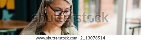 Mature grey woman in earphone working with laptop and papers at cafe indoors