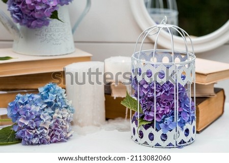 A visual element for the content. Still life in vintage style. Old books, candles, a cage with blue hydrangeas on a white wooden table. A holiday card for the wedding day.