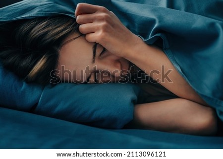 Close-up of the face of a woman who lies in bed, her head is hidden under the covers. Sleepless young woman suffering from insomnia or nightmares. Tired woman closes her eyes with her hands. Royalty-Free Stock Photo #2113096121