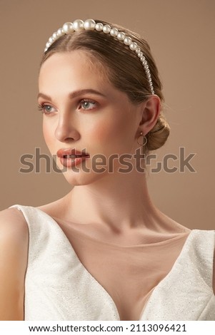 A blonde European girl in a white glitter blouse and golden earrings with charms is posing on the beige background. The girl's hair is fixed with a thin headband adorned w Royalty-Free Stock Photo #2113096421
