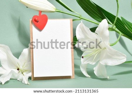 The template is a blank paper and an envelope and a clothespin with a heart with a place for text on a light green background with lily flowers. Flat top view, a place to copy.