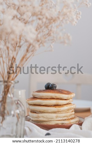 Vertical photo of  stack of pancakes with blueberries and maple syrup. White flowers bokeh style in foreground. Rustic light and airy feel with white light background and copy space available.