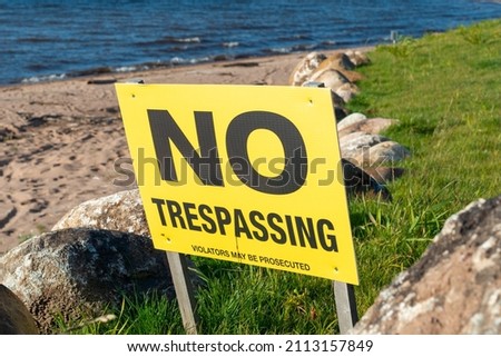 A yellow warning sign with black lettering. The text spells out no trespassing violators will be prosecuted. The private property signpost is on a sandy beach with grass, large rocks and a blue ocean
