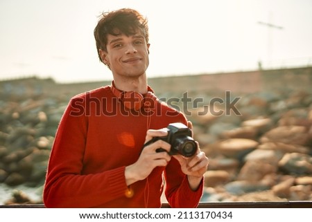 Young hispanic man smiling happy using reflex camera at the beach