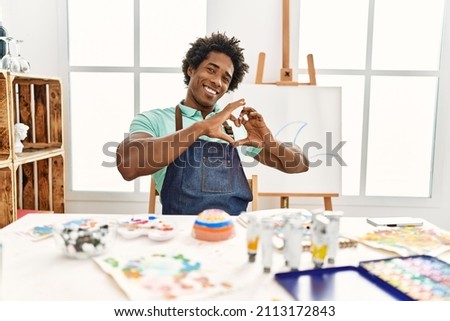 Young african american man sitting on the table at art studio smiling in love showing heart symbol and shape with hands. romantic concept. 
