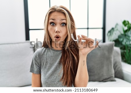Young caucasian woman holding invisible aligner orthodontic scared and amazed with open mouth for surprise, disbelief face 