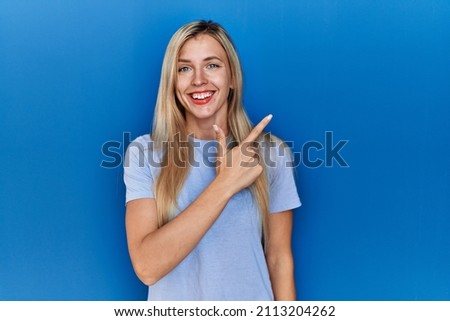 Beautiful blonde woman wearing casual t shirt over blue background cheerful with a smile of face pointing with hand and finger up to the side with happy and natural expression on face 