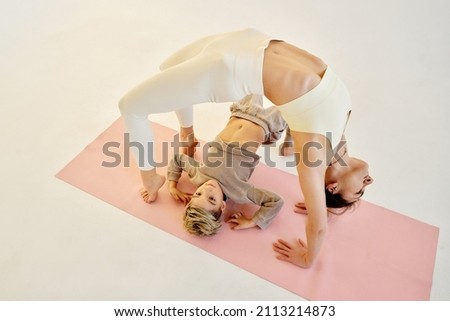 brunette woman with her blonde child son making yoga poses on white background, sunset light in studio