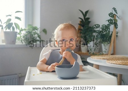 A baby making its first attempts to eat by itself. An infant sitting in highchair in dinning room, holding a spoon and eating Royalty-Free Stock Photo #2113237373
