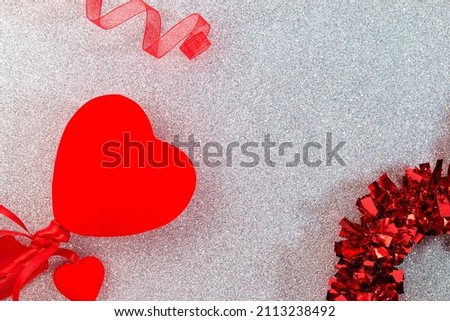 On a silver glitter surface there are red decorations such as a ribbon, hearts and a garland, in the rest of the image there is empty space.