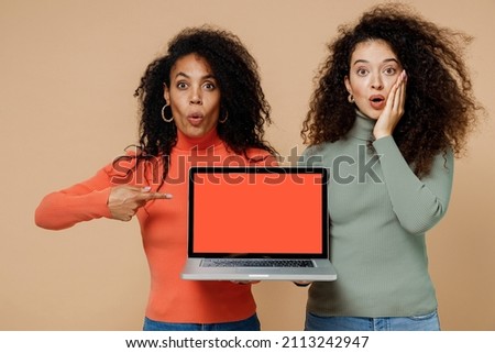 Two shocked young curly black women friends 20s in casual shirts clothes hold use work on laptop pc computer with blank screen workspace area isolated on plain pastel beige background studio portrait
