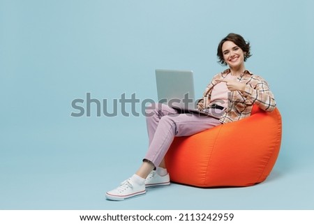 Full body young smiling happy woman 20s in brown shirt sit in bag chair hold use work point finger on laptop pc computer isolated on pastel plain light blue background studio People lifestyle concept