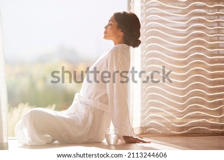Serene woman in bathrobe sitting cross-legged at sunny doorway Royalty-Free Stock Photo #2113244606