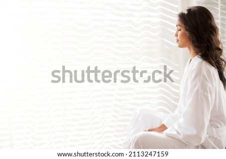 Serene woman in bathrobe meditating at sunny window Royalty-Free Stock Photo #2113247159