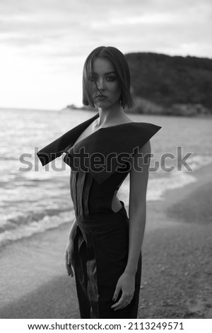 Awesome young woman in fashionable clothes on the sand beach. Beautiful sea landscape on background. Brunette with big eyes and lips. Tan and slim. Summer, sunset. Black and white picture.