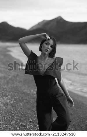 Awesome young woman in fashionable clothes on the sand beach. Beautiful sea landscape on background. Brunette with big eyes and lips. Tan and slim. Summer, sunset. Black and white picture.