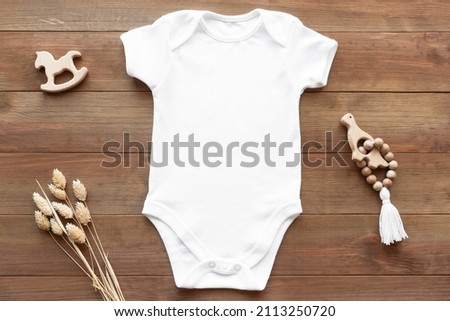 Mockup of white baby bodysuit on dark wood background with dried flowers and toys. Blank baby clothes template, flat lay.
