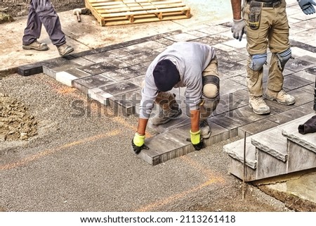 Professional landscape workers paving driveway interlock patio with stone brick at residential construction site. Contractor wearing safety protective cloth on  heavy installation yard work project. Royalty-Free Stock Photo #2113261418