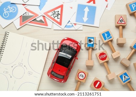 Many different road signs, notebook with sketch of roundabout and toy car on white wooden background, flat lay. Driving school