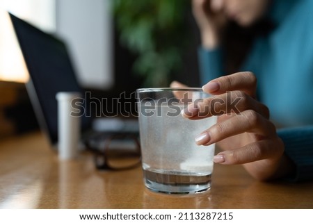 Woman feeling sick in office taking pill medicines, closeup of tablet dissolving in water glass. Ill female holding medication from flu, headache and fever while working at workplace. Selective focus