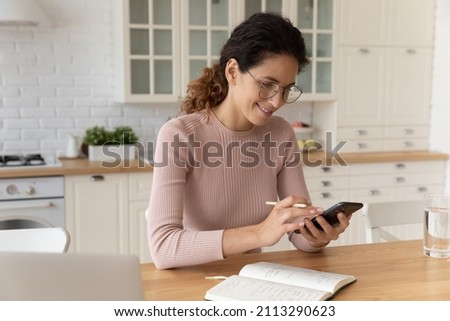 Young hispanic woman in eyeglasses sit at dining table holding smartphone and pencil, check schedule online agenda plan household, surf information on internet writes it in diary, modern tech concept Royalty-Free Stock Photo #2113290623