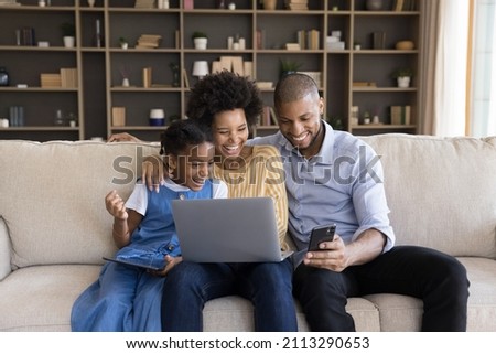 Young parents and little 7s daughter sit on sofa with diverse devices watching on cellphone funny videos laughing looking overjoyed. African family enjoy modern tech, online humorous content concept Royalty-Free Stock Photo #2113290653