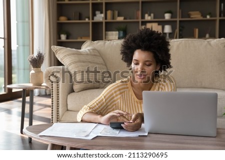 African woman makes calculations on calculator pay utility bills, loan via e-bank application on computer. sit in living room alone. Personal family budget management, modern tech, accounting concept Royalty-Free Stock Photo #2113290695