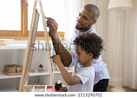 African dad and little son draw on chalkboard with colored chalks. Cute curly haired boy take part at art class with friendly tutor stands by easel enjoy hobby. Family pastime leisure at home concept
