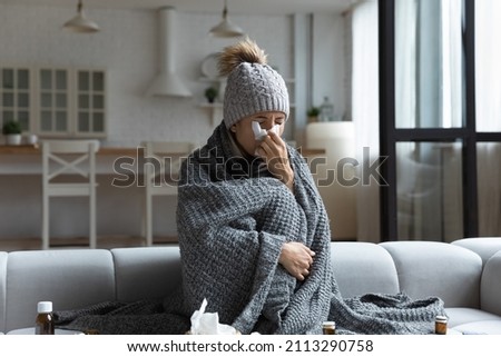 Sick frozen woman sit on sofa covered with knitted plaid, wear hat holds tissue blowing runny nose got fever, catch cold, heap of meds nearby. Seasonal influenza symptoms, flu, unheated flat concept Royalty-Free Stock Photo #2113290758
