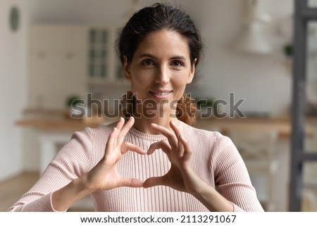 Head shot portrait beautiful loving young Latina woman makes heart symbol sign with joined fingers smile look at camera pose alone at home. Express gratitude, appreciation. I love you gesture concept