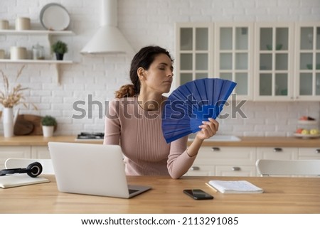 Hispanic woman feel dehydrated has heat stroke, looks overheated cools herself with hand fan, suffers from hot weather, hormonal imbalance rest from work on laptop. Summer, need airconditioner concept Royalty-Free Stock Photo #2113291085