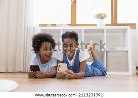 Cheerful little African siblings lying on warm floor at home with smartphones, spend leisure on internet, having fun using amusing mobile application. Generation z, device overuse, modern tech concept