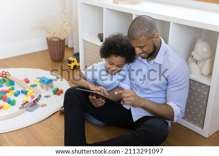 African dad interested little son using digital tablet sit on warm floor in cozy nursery. Family spend weekend having fun on internet, enjoy new cool online game. Modern tech, leisure at home concept