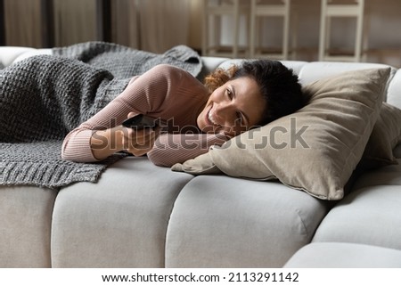 Hispanic young smiling woman lying on couch cushion holds TV remote controller switch channels watch internet television, single 30s female enjoy day off at cozy home. Lifestyle, lazy holidays concept Royalty-Free Stock Photo #2113291142