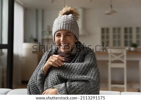 Funny laughing Hispanic woman wear knitted grey bobble hat wrapped in warm plaid sit on sofa relaxing warmup while watch comedy TV show, spend carefree weekend alone at home having fun feel overjoyed Royalty-Free Stock Photo #2113291175