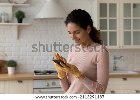 Latina housewife woman in protective latex gloves distracted from housework cleaning kitchen using smartphone, text share message, search on internet professional cleanup products, lifestyle concept