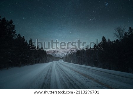 A night road and beautiful scenery with starry night sky. Winter landscape with empty road, forest and mountains. 