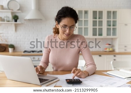 Hispanic woman use calculator calculate costs working sit at table in kitchen. Pay monthly bills through e-bank app on laptop. Business, summarize household expenses, manage budget, accounting concept Royalty-Free Stock Photo #2113299905