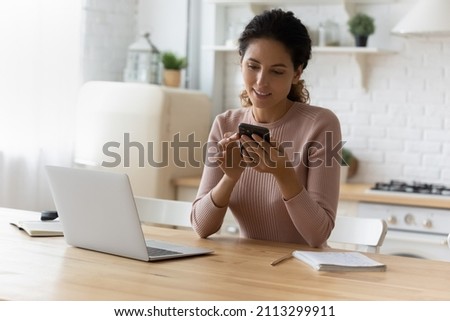 Attractive 30s Latin woman sit at table with laptop in kitchen using smartphone. Make easy remote food delivery order through mobile application, scroll news feed in social media, modern tech concept