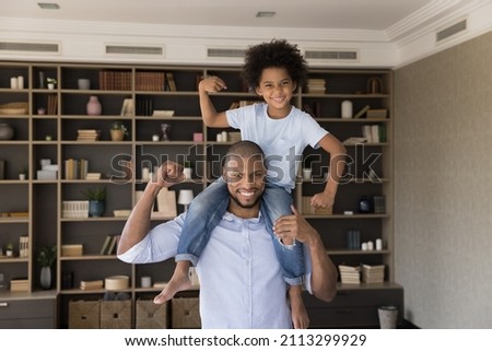 Happy African family showing strength of their arms at camera, little 6s cute son sit on handsome dad shoulder demonstrate muscles of their biceps. Concept of healthy sporty lifestyle, workout at home Royalty-Free Stock Photo #2113299929