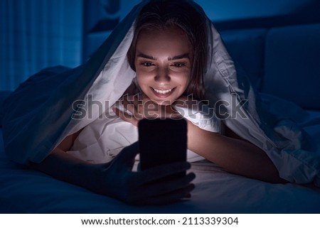 Content female lying in bed under blanket and watching movie on smartphone in dark bedroom before sleep in late evening Royalty-Free Stock Photo #2113339304